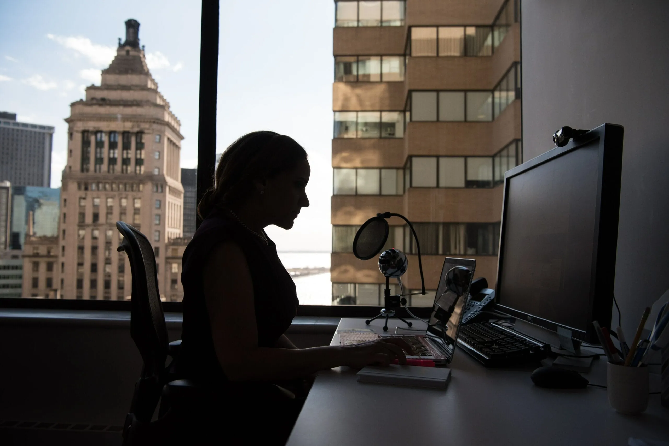 Silhouette einer Frau, die am Schreibtisch vor einem großen Bürofenster mit Blick auf Hochhäuser arbeitet