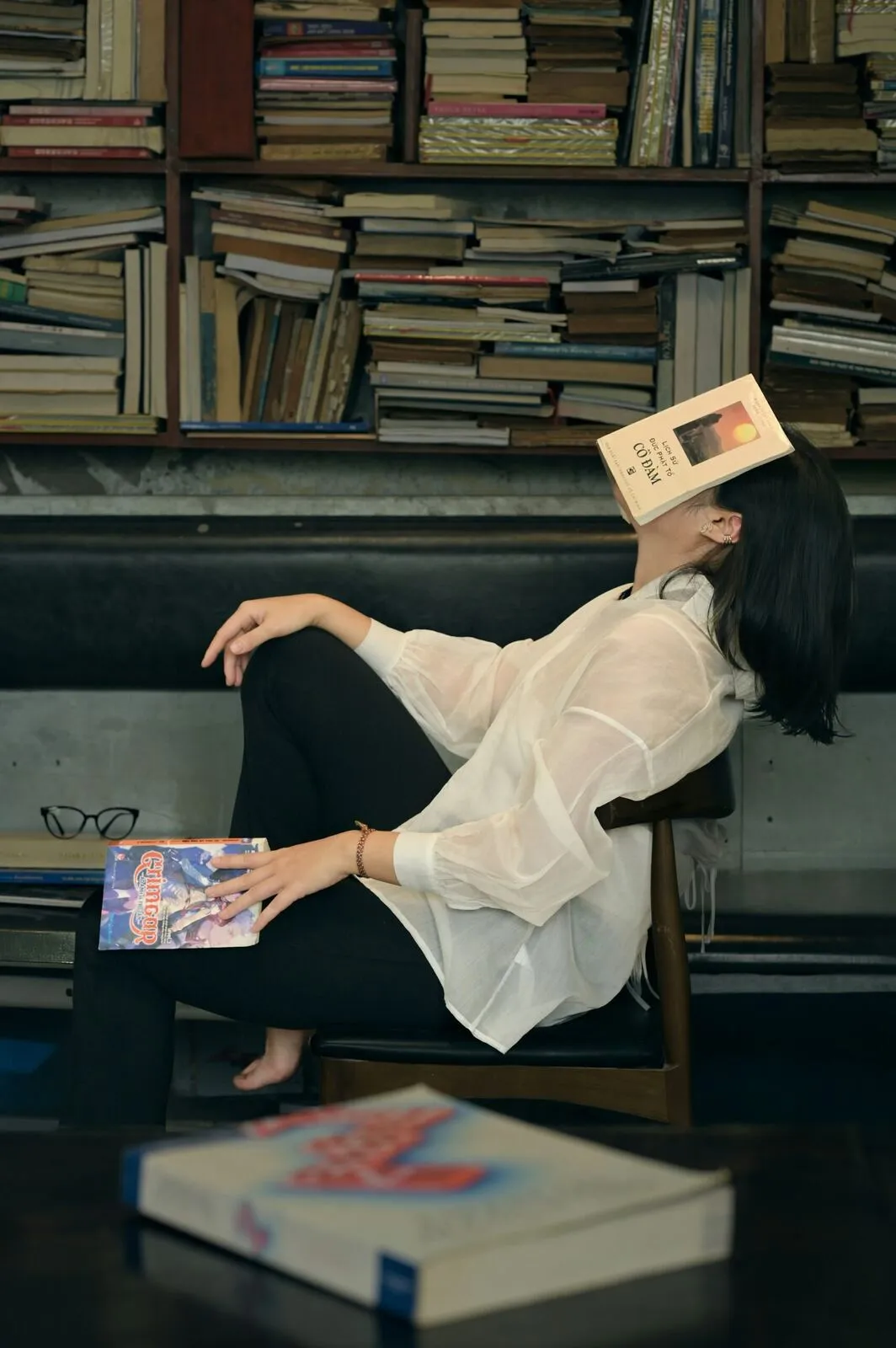Eine Frau lehnt erschöpft im Sessel, ein aufgeschlagenes Buch über dem Gesicht, umgeben von Bücherstapeln