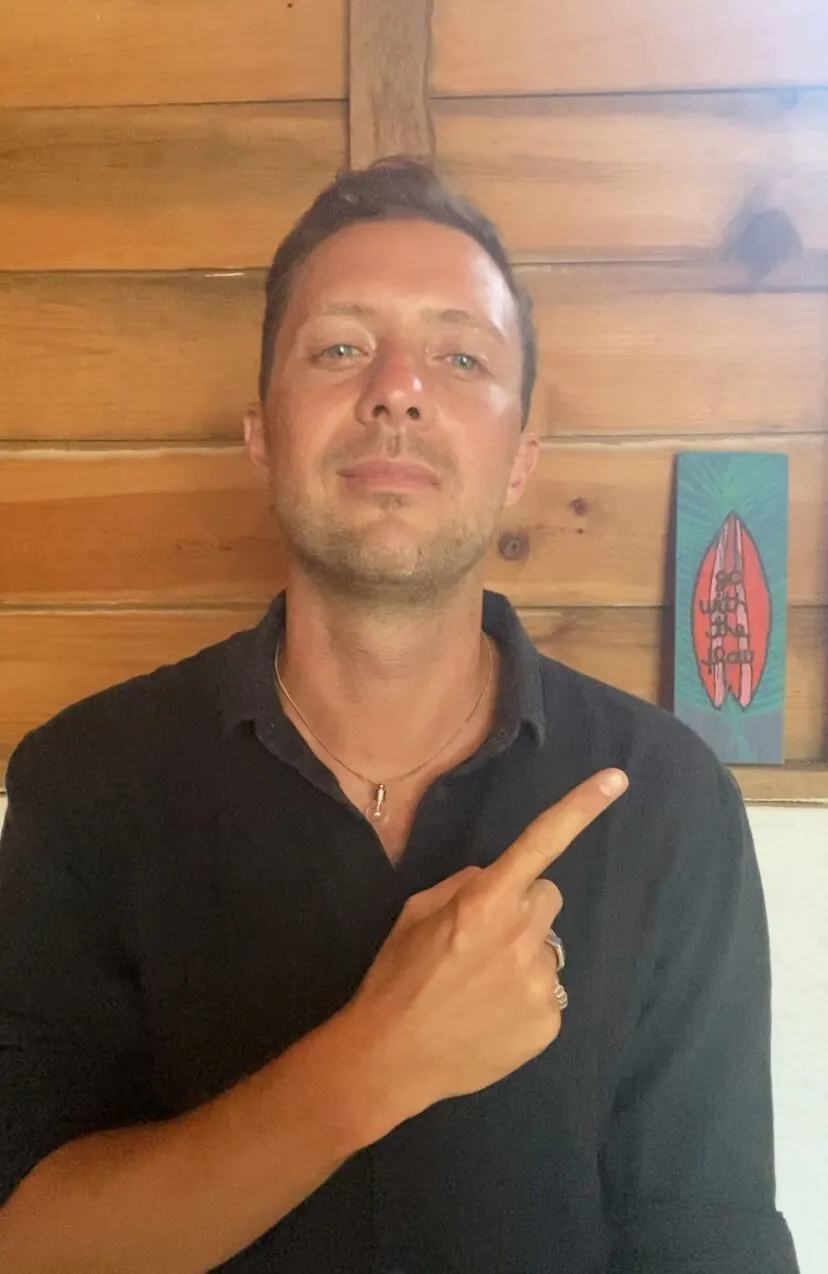 Simon Karim vor einer Holzwand, zeigt lächelnd auf sich